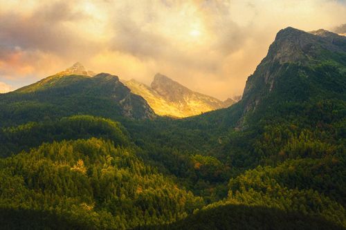Free Dramatic mountain landscape with sunlit peaks and lush green forests beneath a cloudy sky. Stock Photo