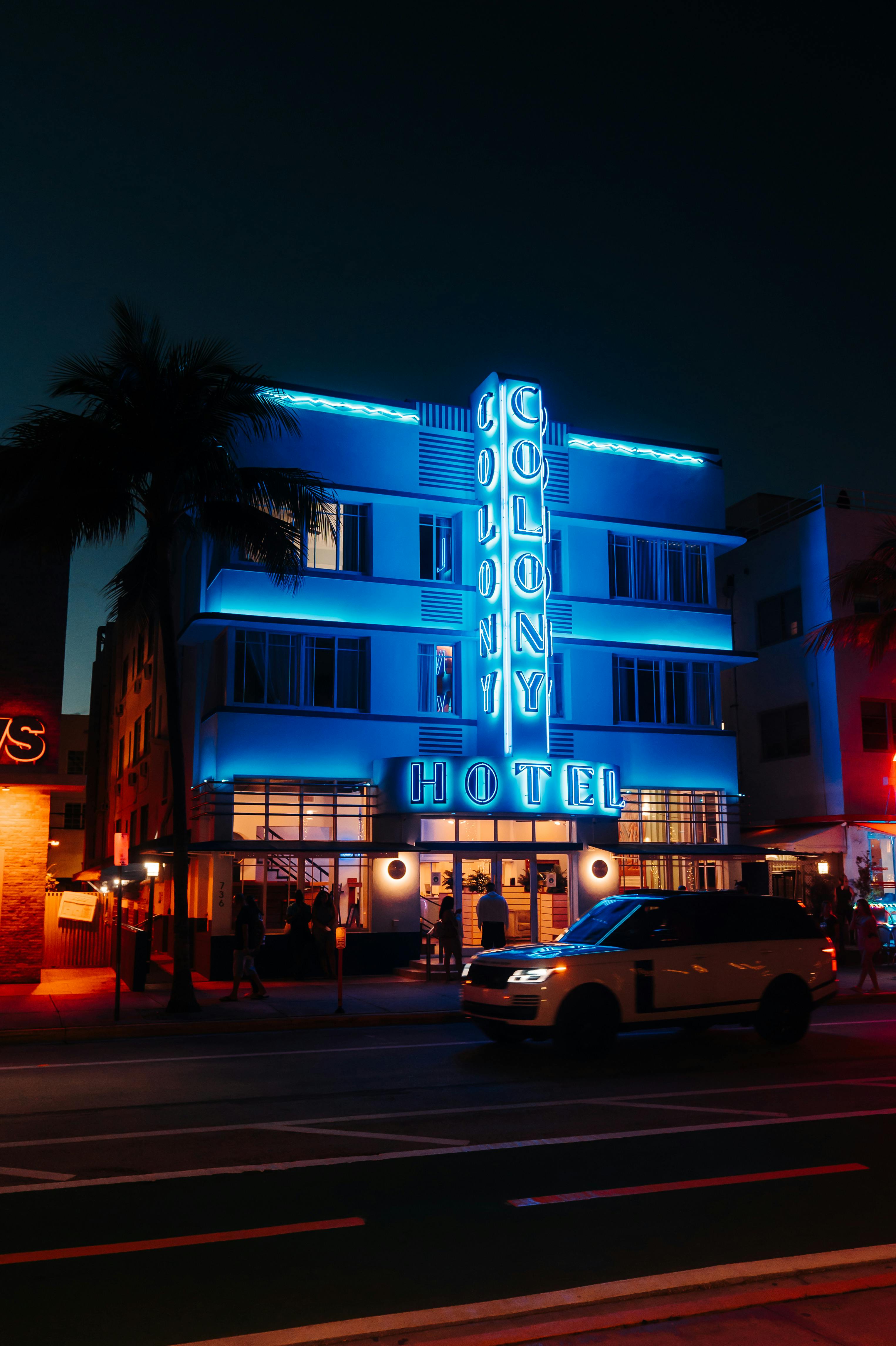 Illuminated Colony Hotel with vibrant neon lights against a night sky and a classic car parked nearby.