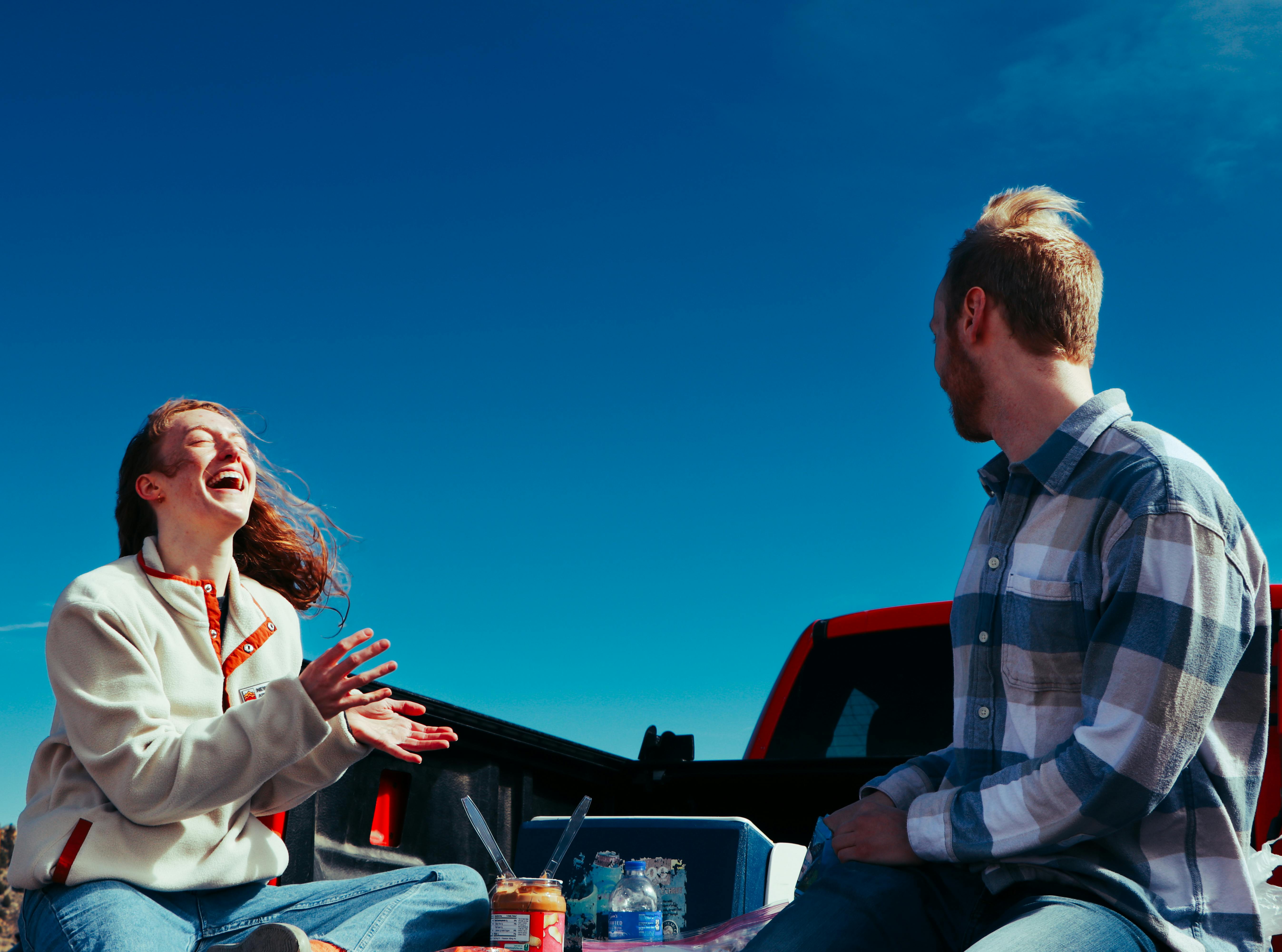 Two friends share a joyful moment outdoors, sitting by a truck under a clear blue sky.