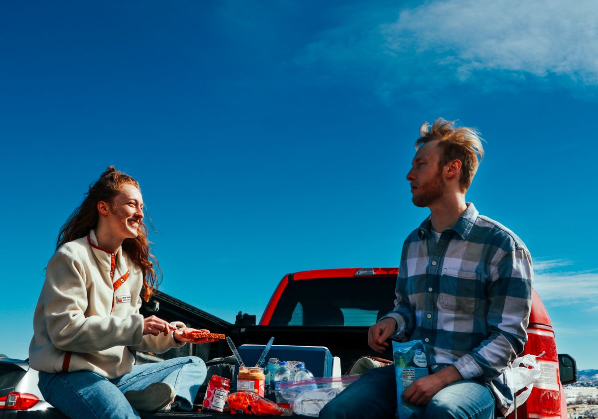 Couple relaxing on the open tailgate of a red pickup truck with a cooler and snacks on game day