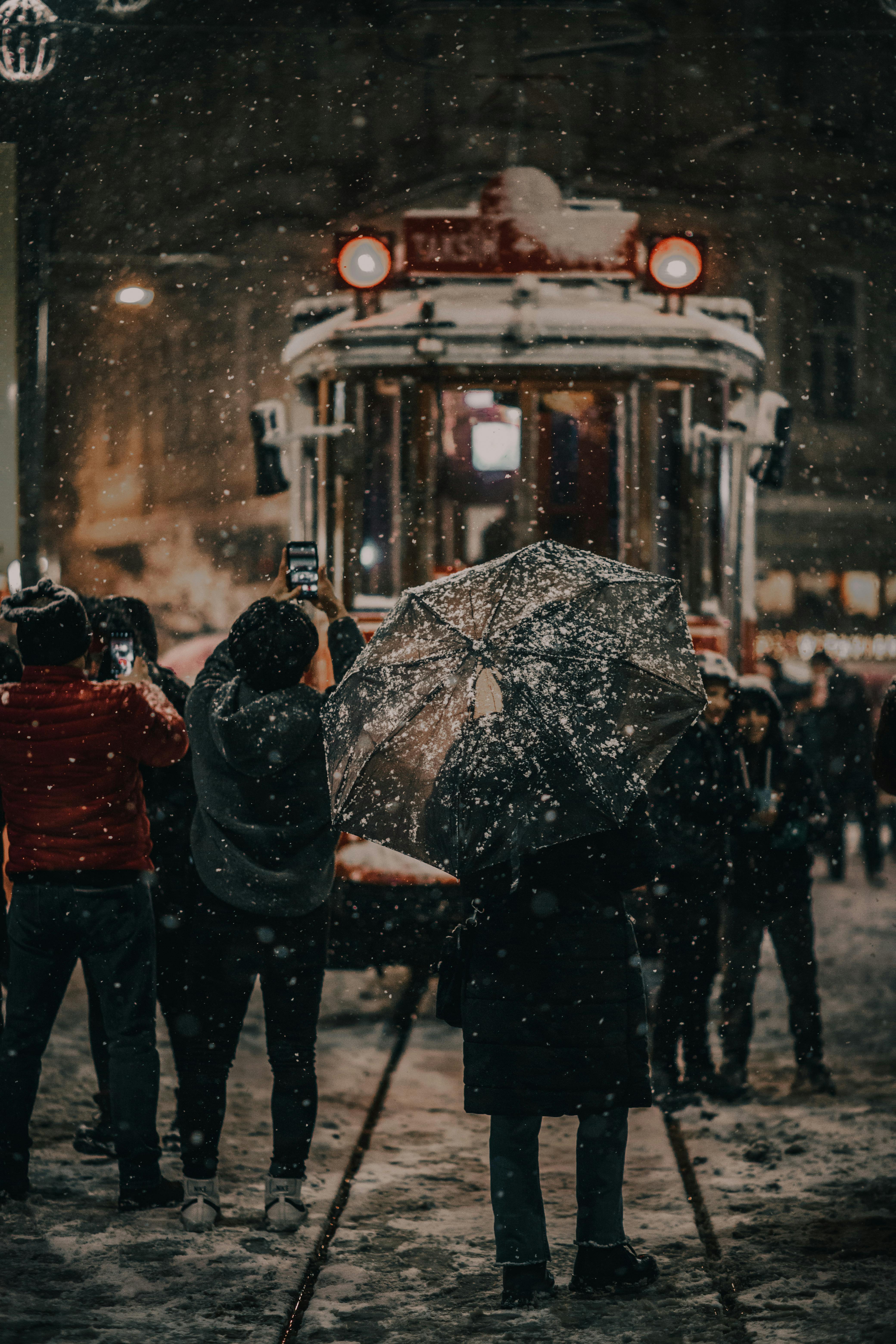 Snowy night urban scene with people, tram, and umbrella in winter cityscape.