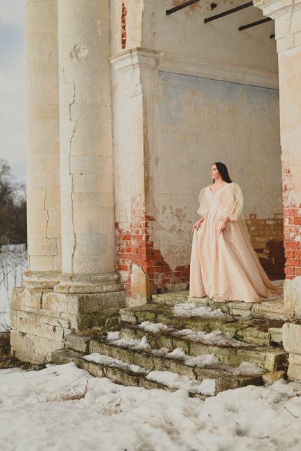 A woman in a vintage gown stands on snowy steps, framed by rustic architecture, presenting a winter fashion scene.