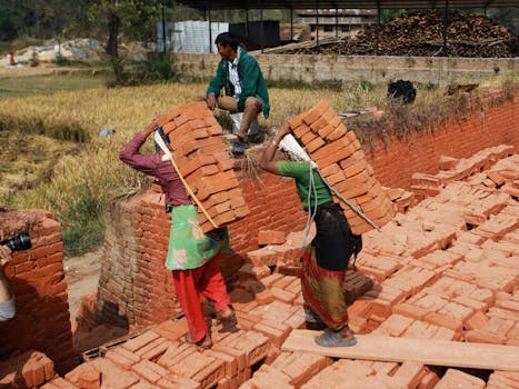 Two laborers carrying red bricks in an outdoor factory setting. Manual work and industry.