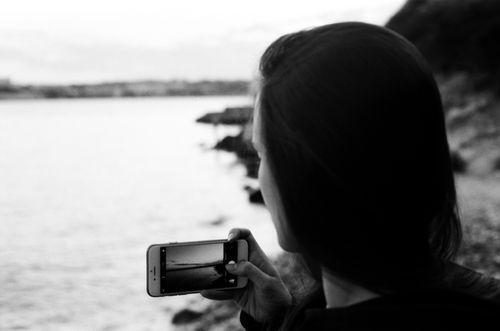 Free Black and white photo of a woman capturing the ocean view with her smartphone on a serene day. Stock Photo