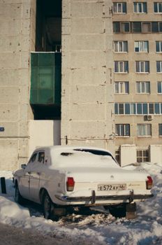 Classic car dusted with snow parked beside tall urban structure.