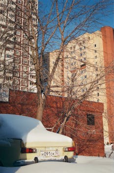 Classic car in snow with leafless trees and urban buildings during winter.
