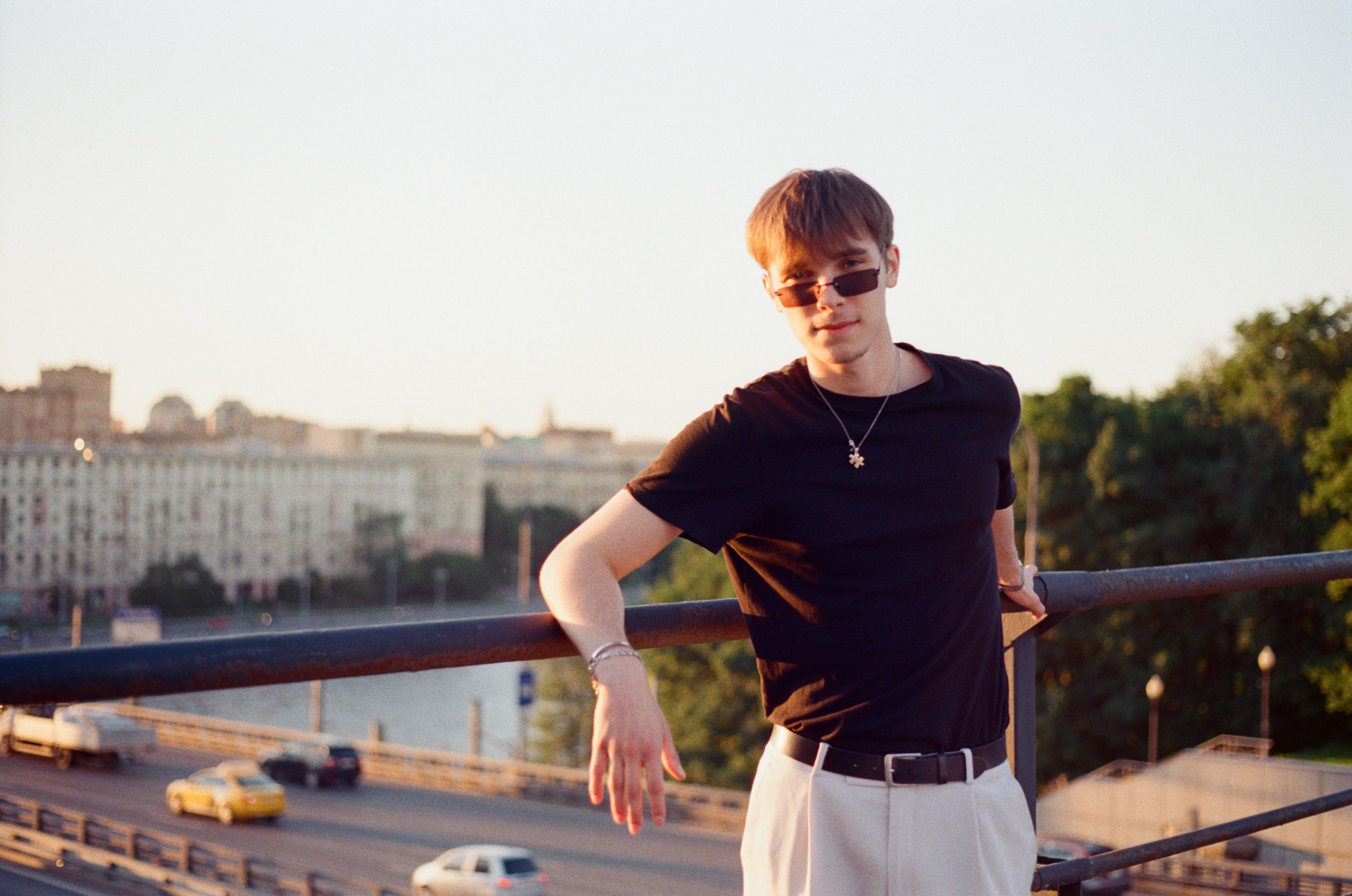 Stylish young man in sunglasses leaning against a railing with an urban backdrop in warm sunset light.