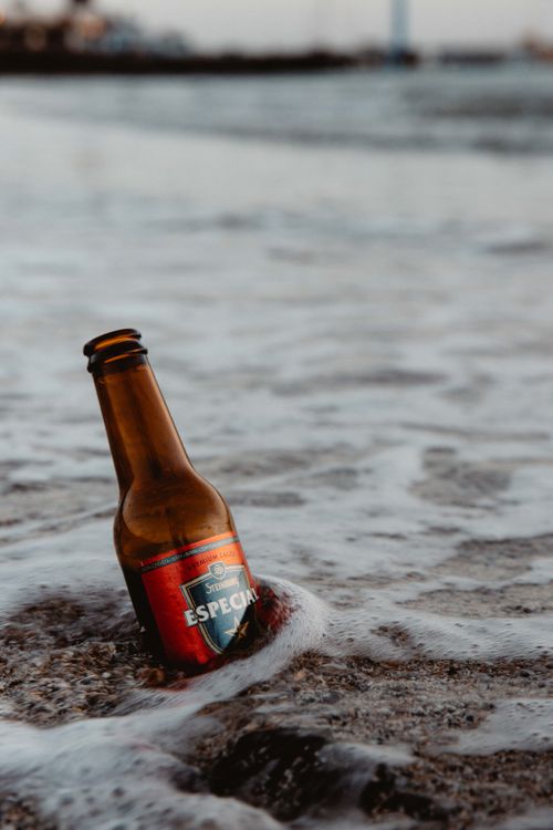 Free Close-up of an empty beer bottle on the sandy beach with ocean waves. Stock Photo