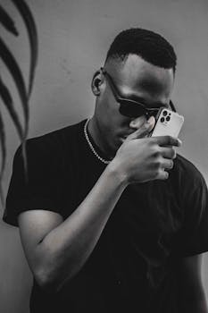 A fashionable man in sunglasses holds a smartphone in a grayscale portrait setting.