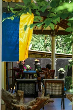 A child sits on a terrace with a tablet during a sunny summer day, surrounded by greenery and vibrant colors.