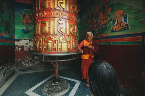 Free A senior monk standing beside a large ornate prayer wheel inside a colorful temple. Stock Photo