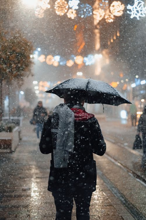 Free Back view of a man with an umbrella walking in a snowy city night, festive lights overhead. Stock Photo