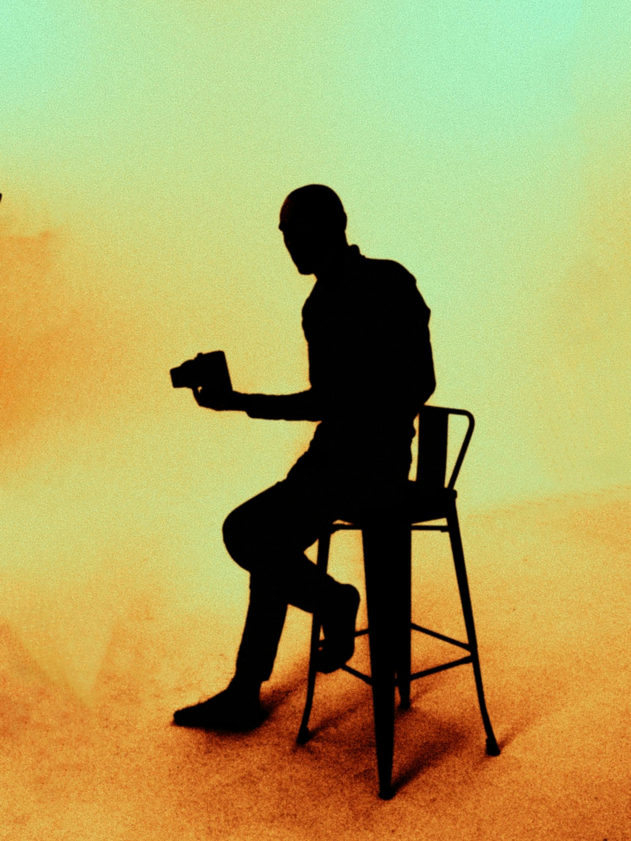 Silhouette of man sitting on a chair holding an object against a warm gradient background.
