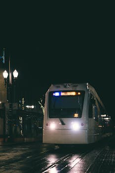 A tram travels through an illuminated city street at night, reflecting on wet pavement.