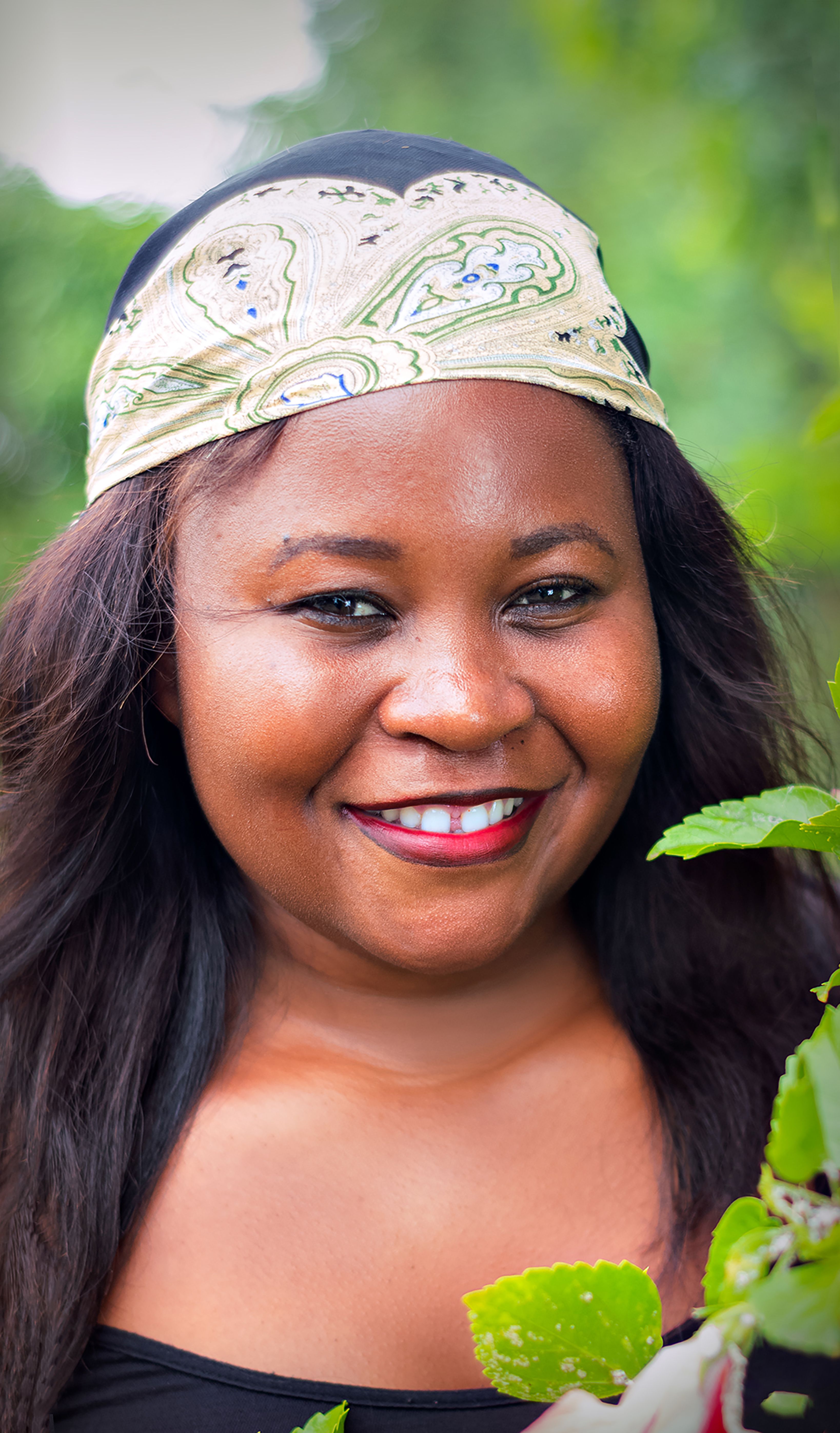 Portrait of a smiling woman wearing a headscarf, surrounded by greenery, exuding joy and serenity.