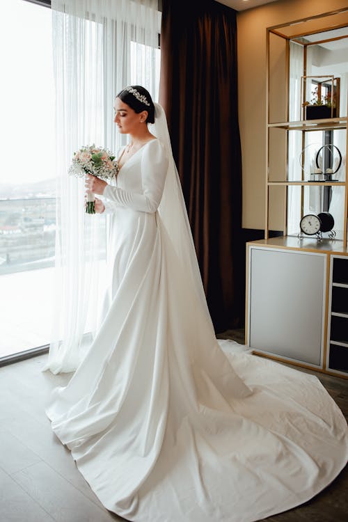 Woman in White Wedding Dress Standing Beside Window