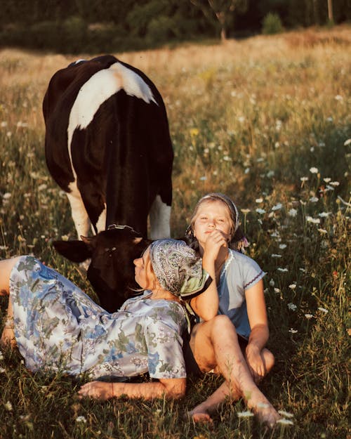 Mother and Daughter Sitting in Meadow Next to Grazing Cow