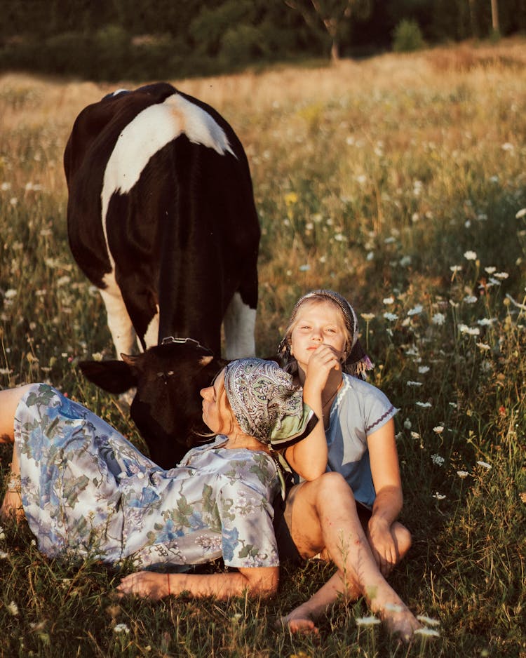 Mother And Daughter Sitting In Meadow Next To Grazing Cow