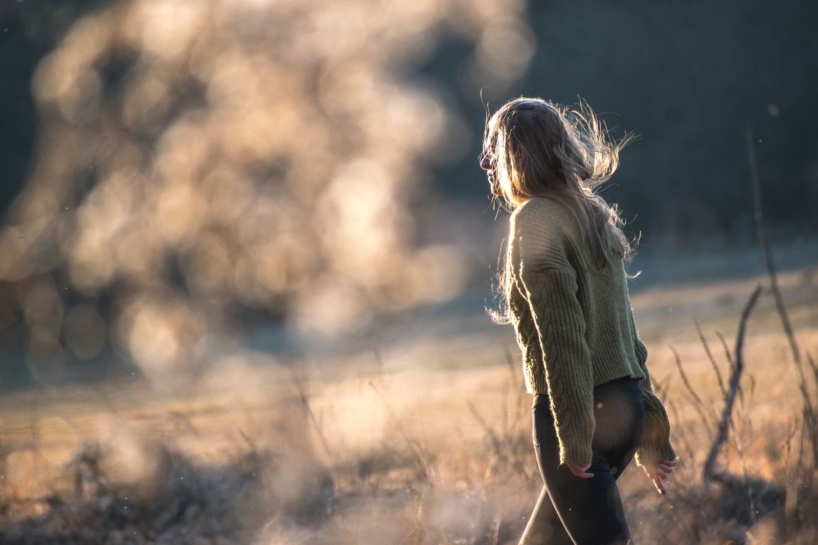 Woman in an olive sweater walking through a sunlit golden field, hair lifted by the breeze