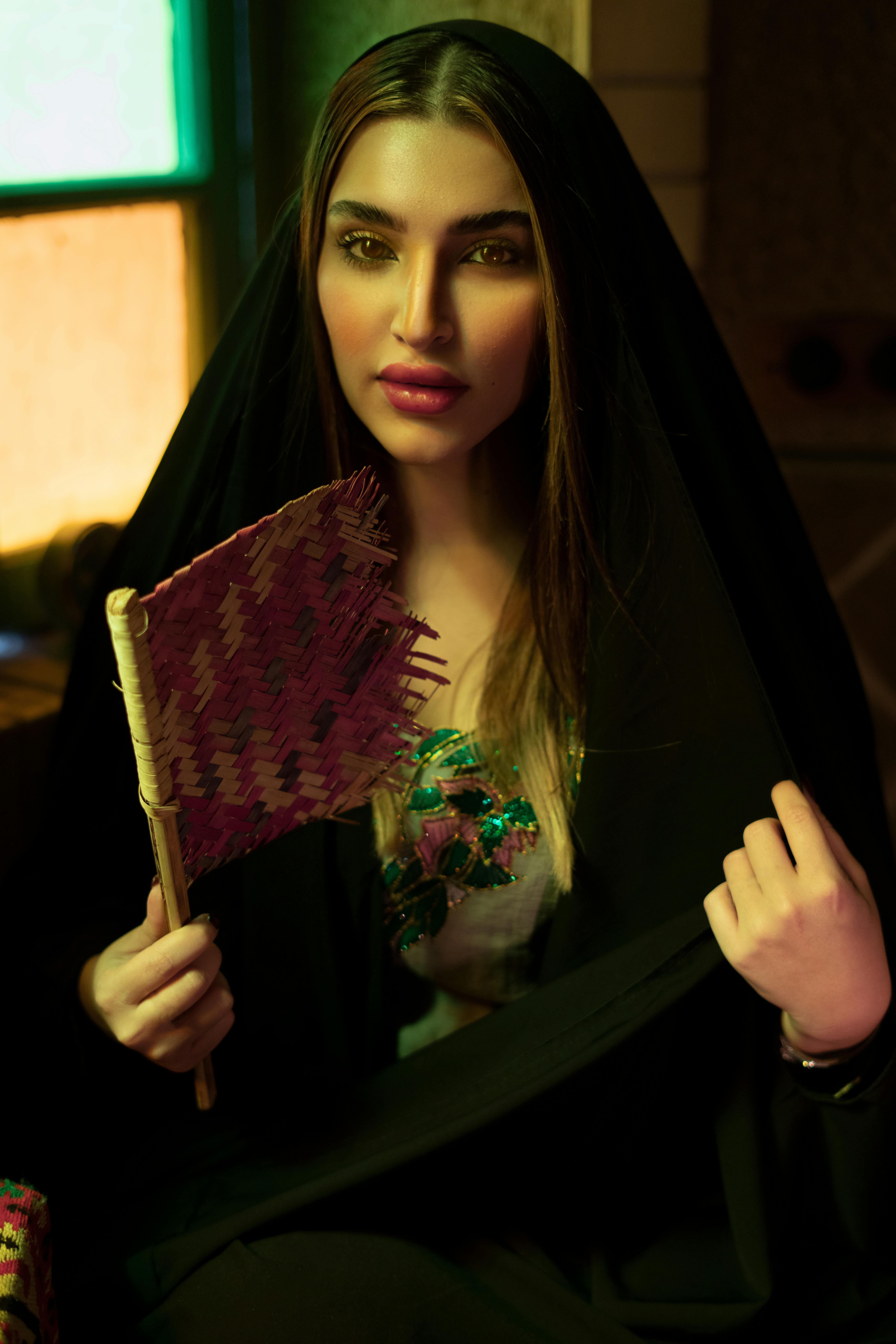 Portrait of a woman in a headscarf holding a traditional fan indoors in warm lighting.