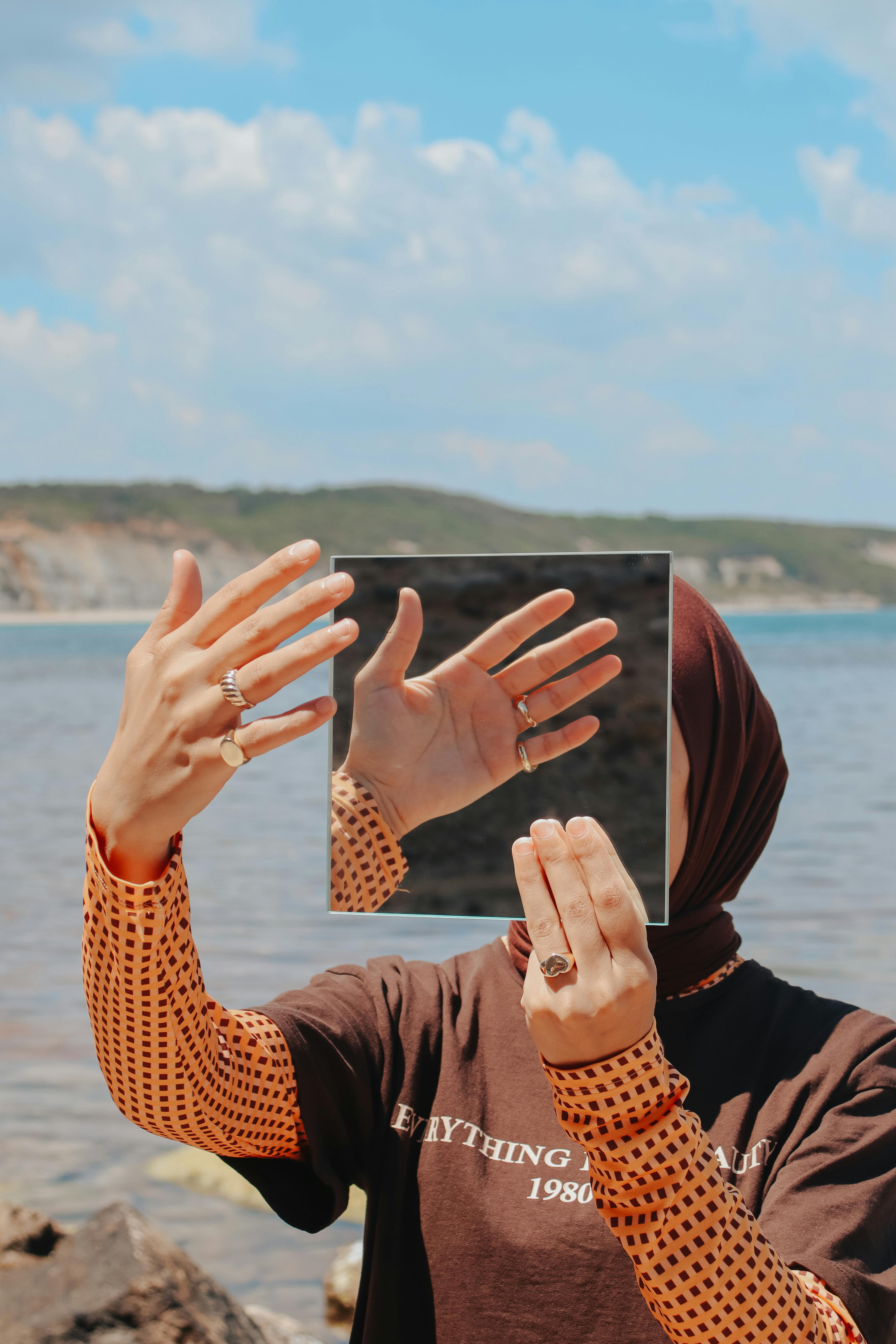 A woman holding a square mirror reflecting her hands by the sea on a sunny day.