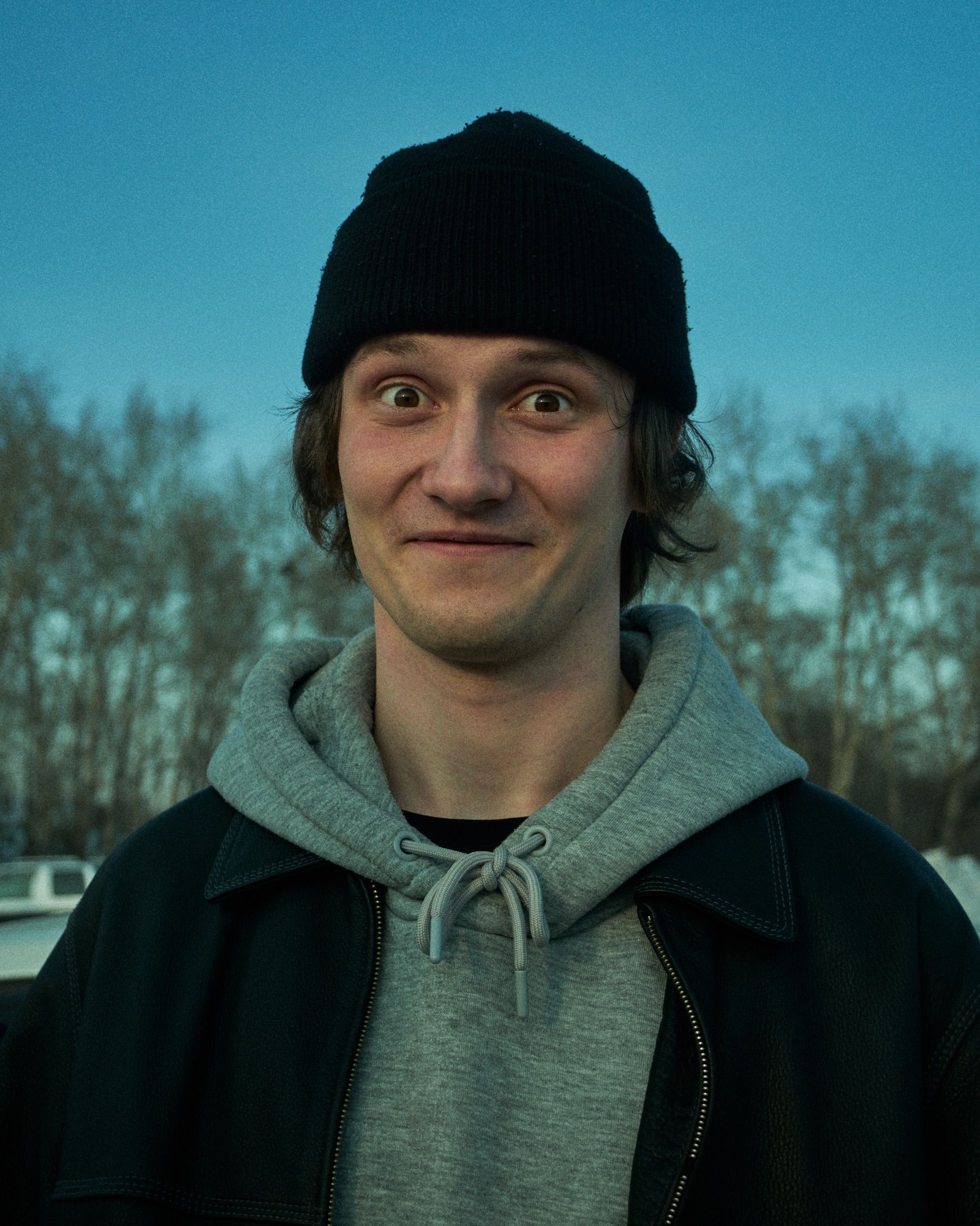 A man wearing a beanie and hoodie smiles outdoors in a winter setting.