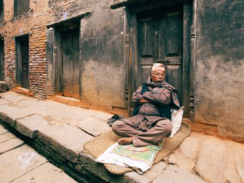 Free An elderly man sits peacefully on a sidewalk in a historic street, embracing tradition and calm. Stock Photo