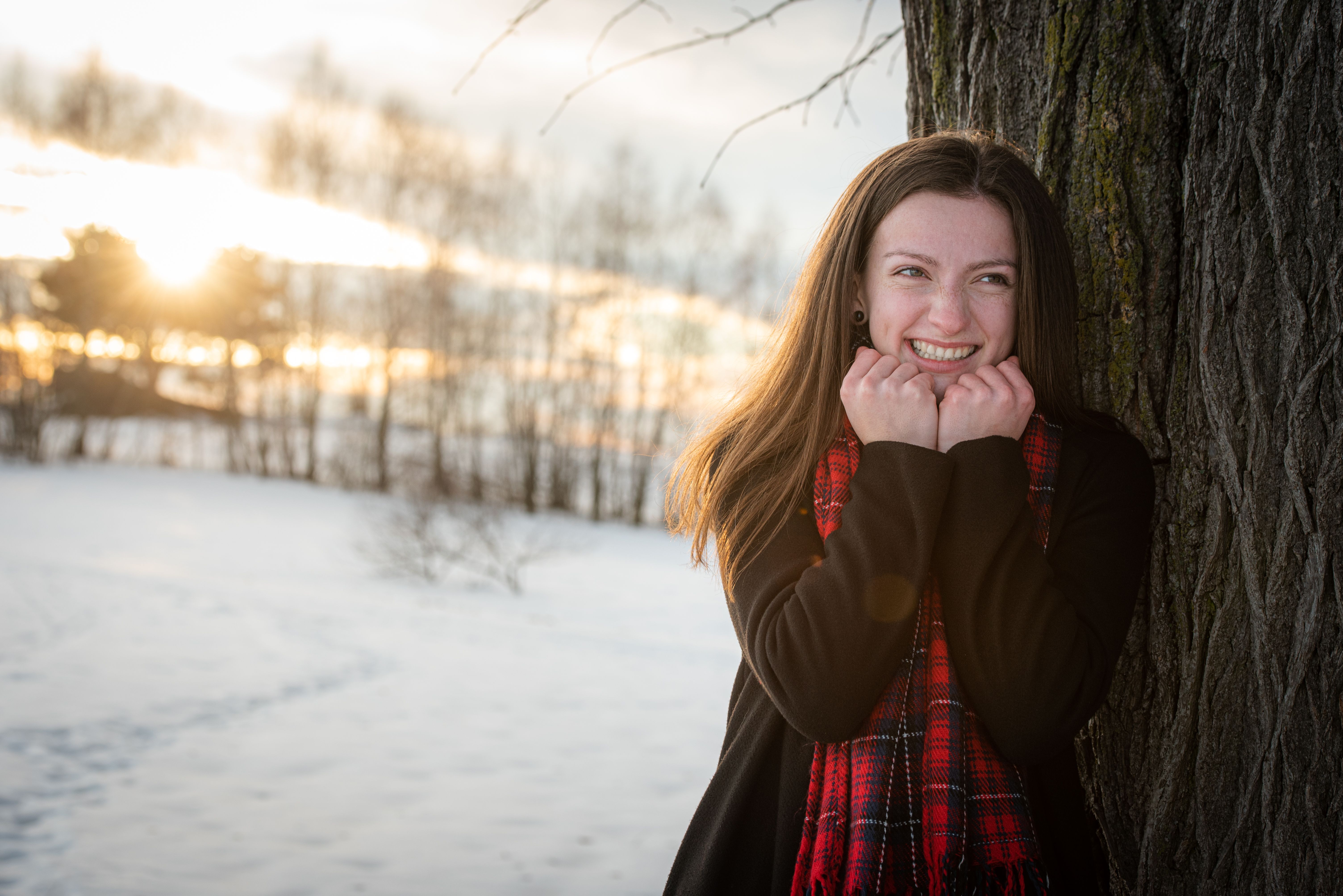 Happy woman in winter attire leaning against a tree, smiling at sunset.