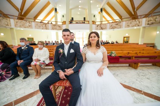A bride and groom seated in a beautifully decorated church, surrounded by guests during their wedding ceremony.