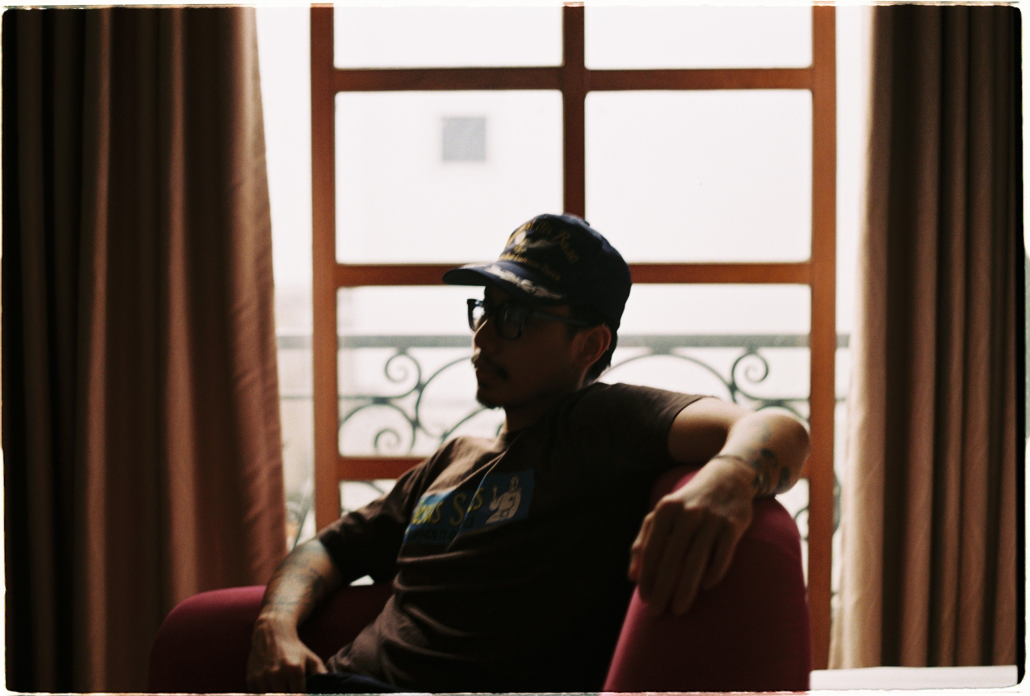 A man wearing a cap relaxes in a cozy armchair, indoors by a window.