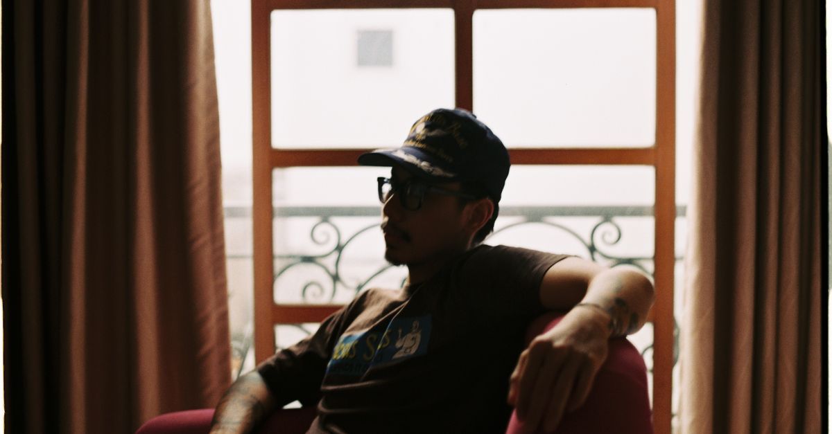 A man wearing a cap relaxes in a cozy armchair, indoors by a window.