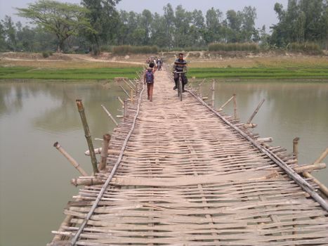 A rustic bamboo footbridge crossing a calm river with people walking and a man bicycling.