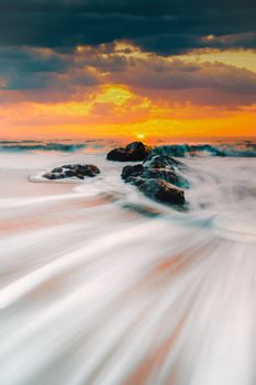 Stunning long exposure photograph capturing waves flowing over rocks during a vibrant sunset at the coast.