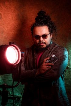 A stylish man with curly hair and sunglasses poses with a camera under dramatic lighting.