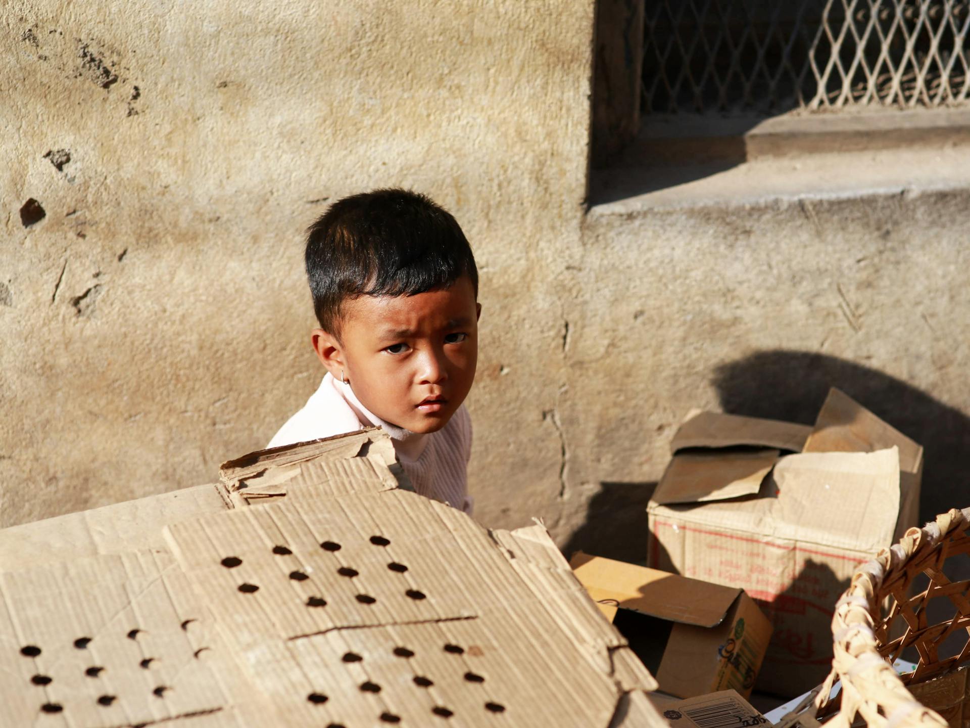 A young boy glances curiously while surrounded by cardboard boxes outdoors in bright sunlight.