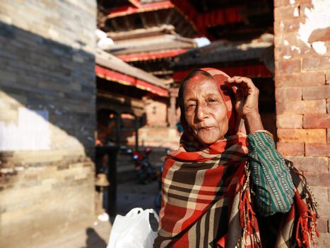 An elderly woman stands in sunlight wearing a traditional shawl against a backdrop of historic architecture.