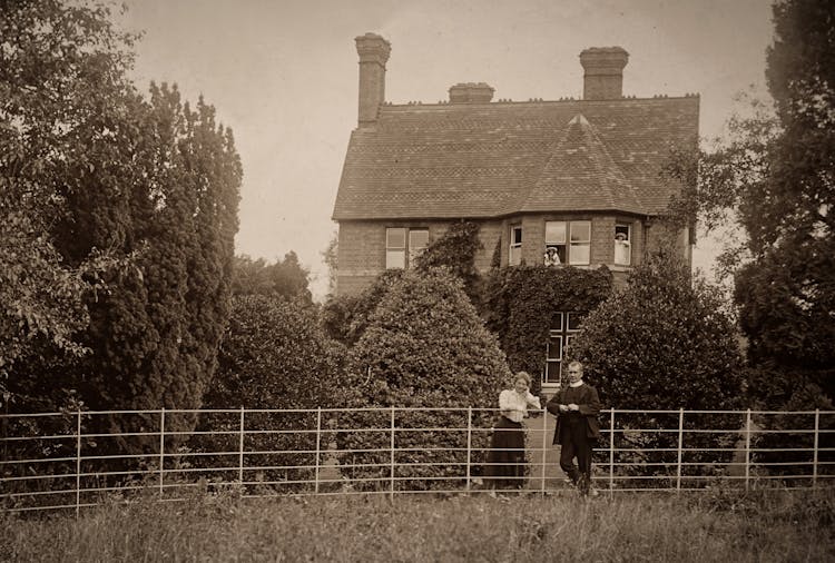 Country House And Garden With Man And Woman Standing Near A Fence