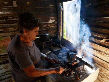 An elderly woman cooking on a rustic wooden stove inside a cabin, with natural light streaming in.