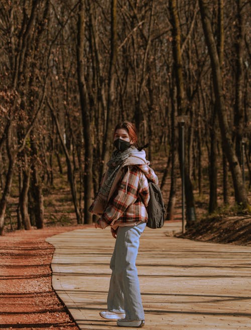 A Woman in Brown and White Plaid Scarf Walking on Pathway in Between Trees