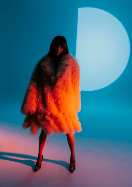 Fashion model in a fur coat stands dramatically lit in a studio with a blue backdrop.