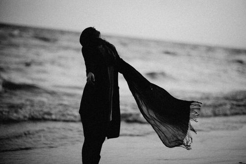 Free An artistic black and white silhouette of a woman standing by the ocean shore with a flowing scarf. Stock Photo