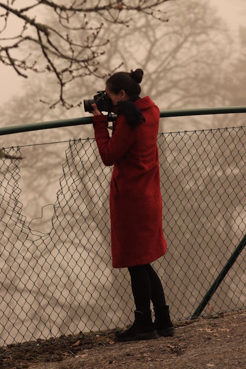 A Woman in Red Coat Holding a Black Dslr Camera Standing Beside Metal Fence