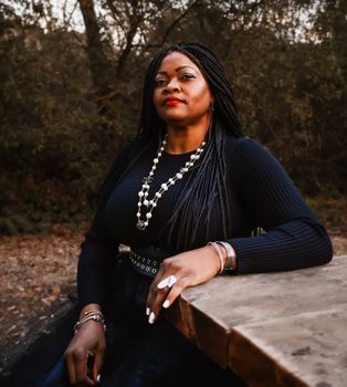 Stylish African woman with braided hair, pearl necklace, and bracelets sitting elegantly in a park.