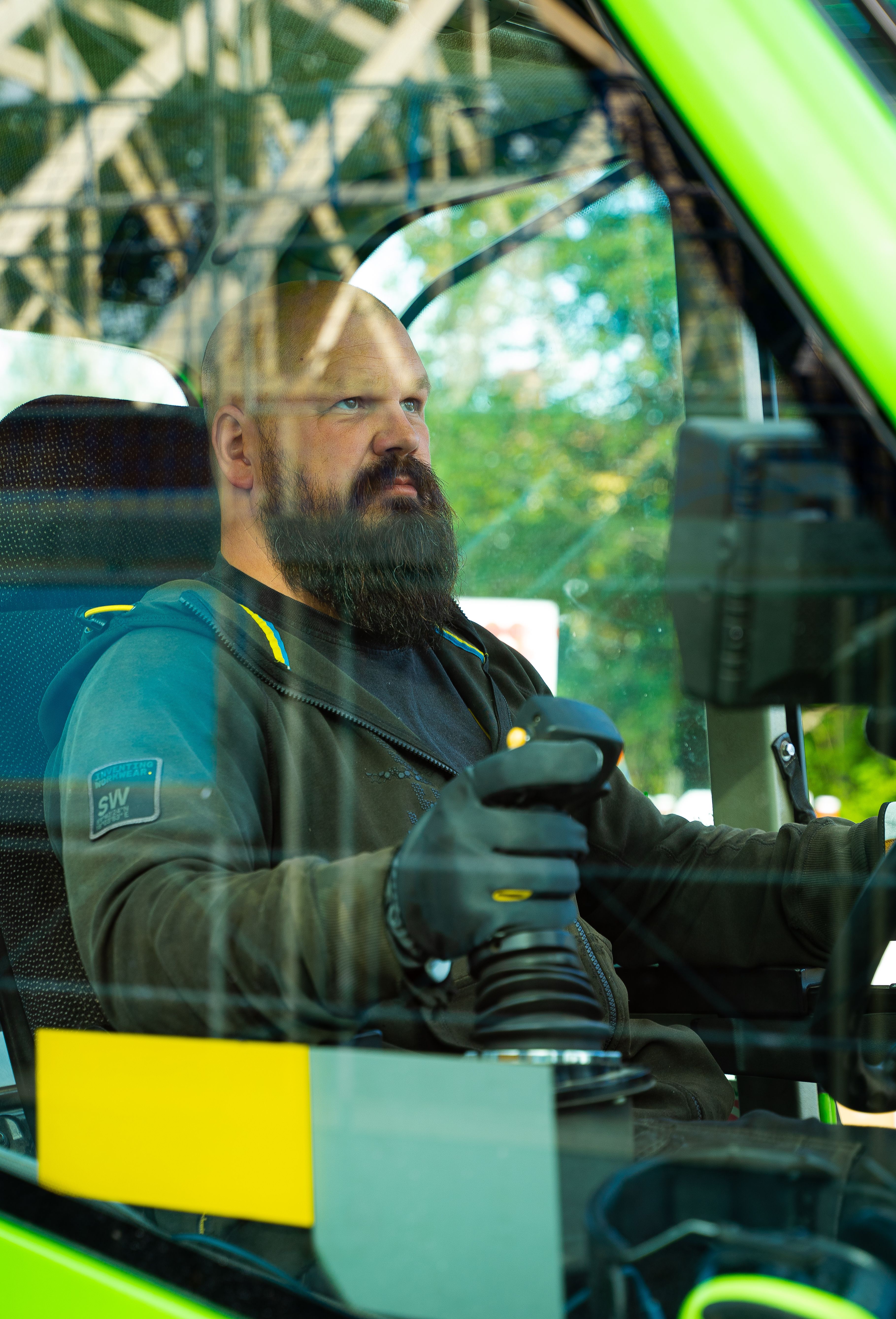 Bald bearded man using controls inside machinery cab, seen through glass window.
