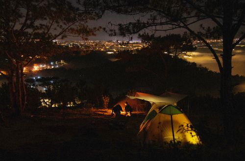 Free A serene camping scene at night with tents and a bonfire under a night sky and city lights in the distance. Stock Photo