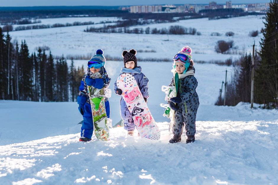 Three children holding snowboards in winter gear on a snowy slope, captured in bright daylight.