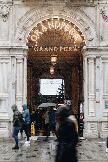 Busy entrance at Grand Pera, Istanbul, featuring people in motion on a rainy day.