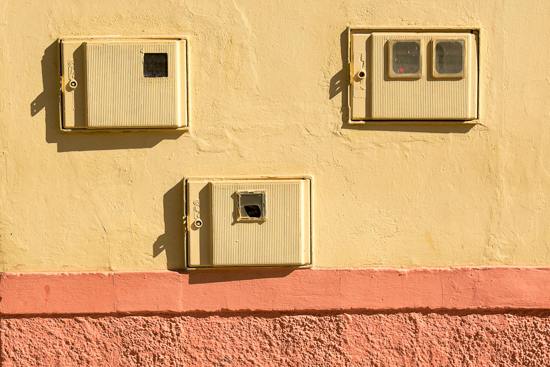 Close-up of vintage electrical boxes on a yellow wall with sunlight casting shadows.