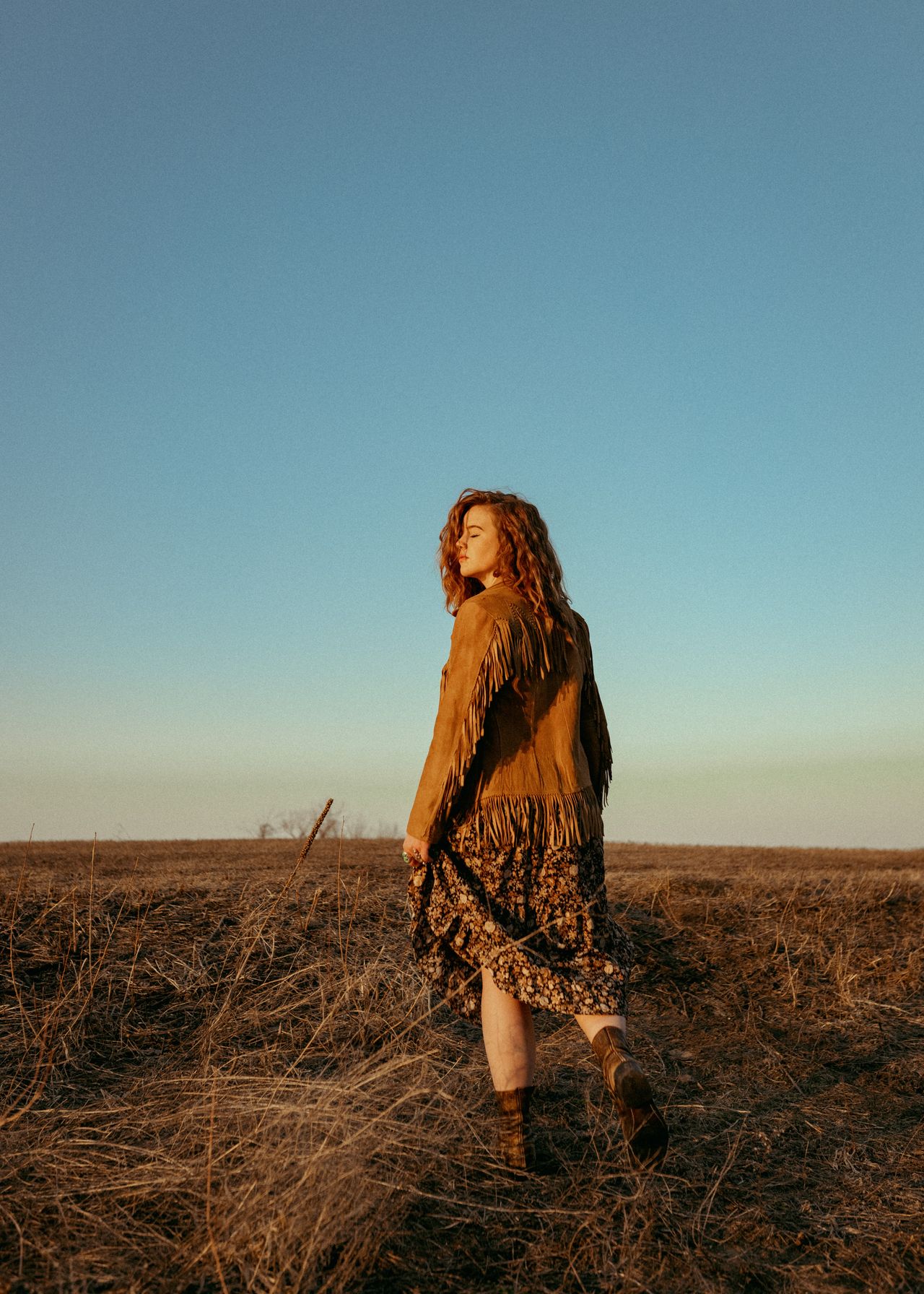 Woman in a tan suede fringe jacket and leopard skirt standing in a field
