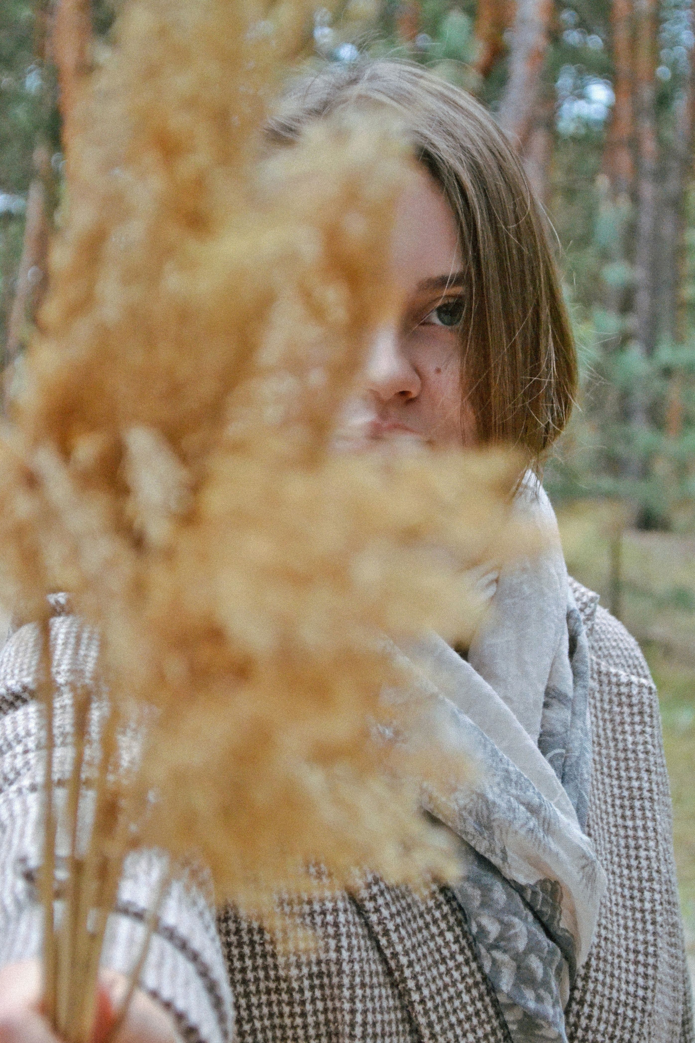 A close-up portrait of a woman holding yellow grass outdoors in a forest.